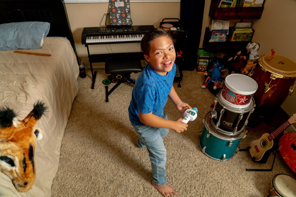 An overhead view of a child wearing a blue shirt and light blue jeans as they stand in the middle of their bedroom surrounded by toys. The child holds a microphone toy with their hands.